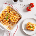 Baked lasagna in a white baking dish and a serving on a plate, garnished with basil, with tomatoes and cutlery nearby.