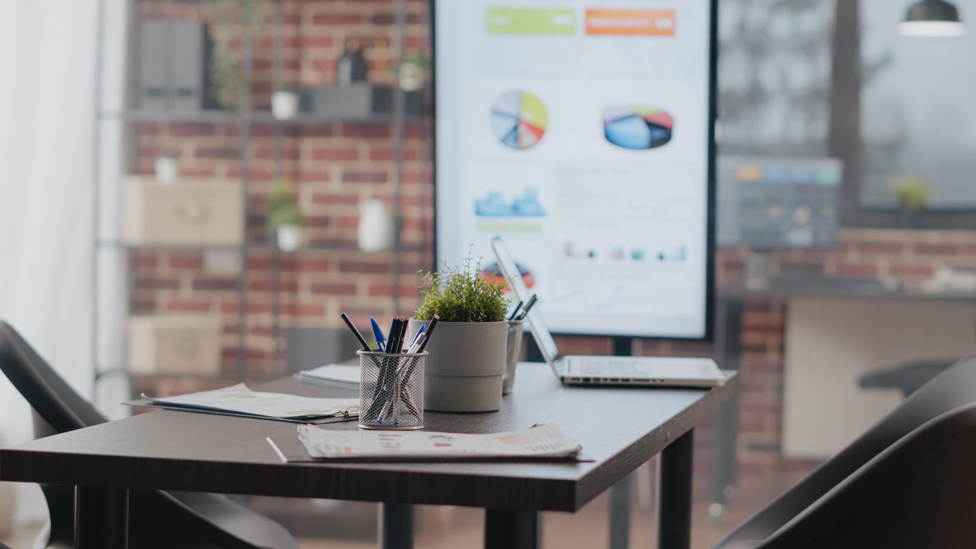 A modern office desk with pens, a small potted plant, and a laptop, facing a display screen showing business data charts.