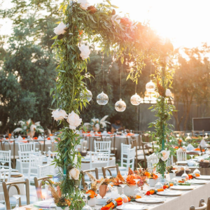 An outdoor wedding reception with long tables, floral centerpieces, hanging ornaments, and greenery under sunset light.