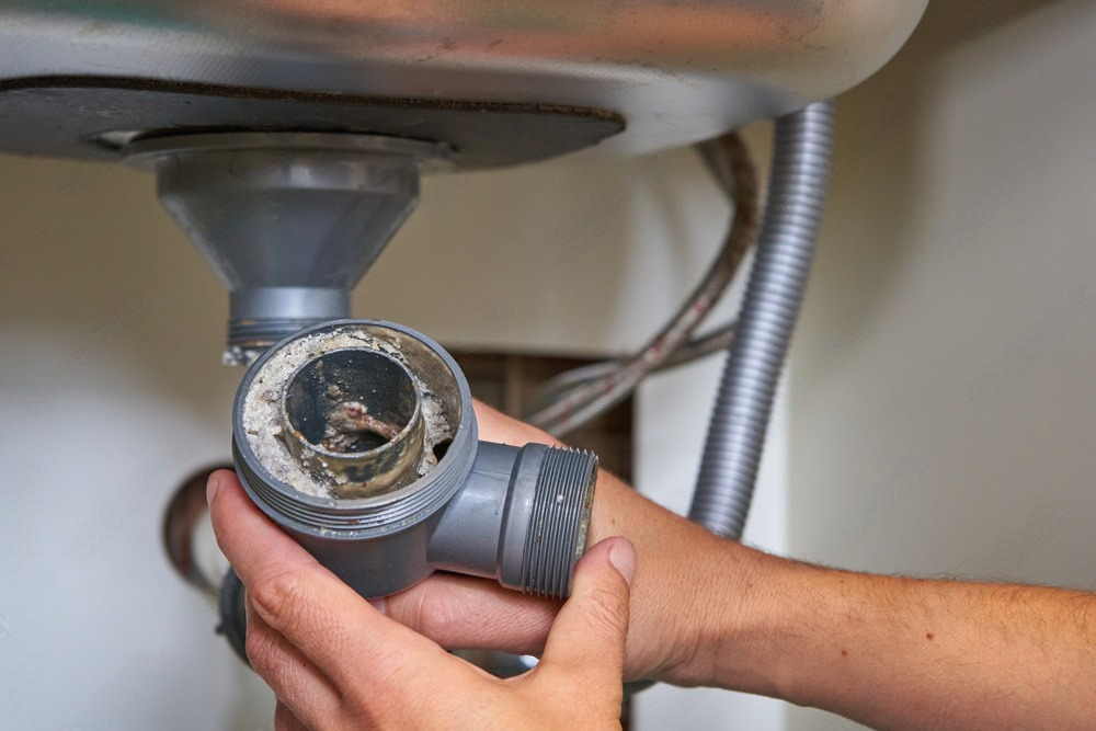 A hand holds a dirty sink drain pipe under a kitchen sink.