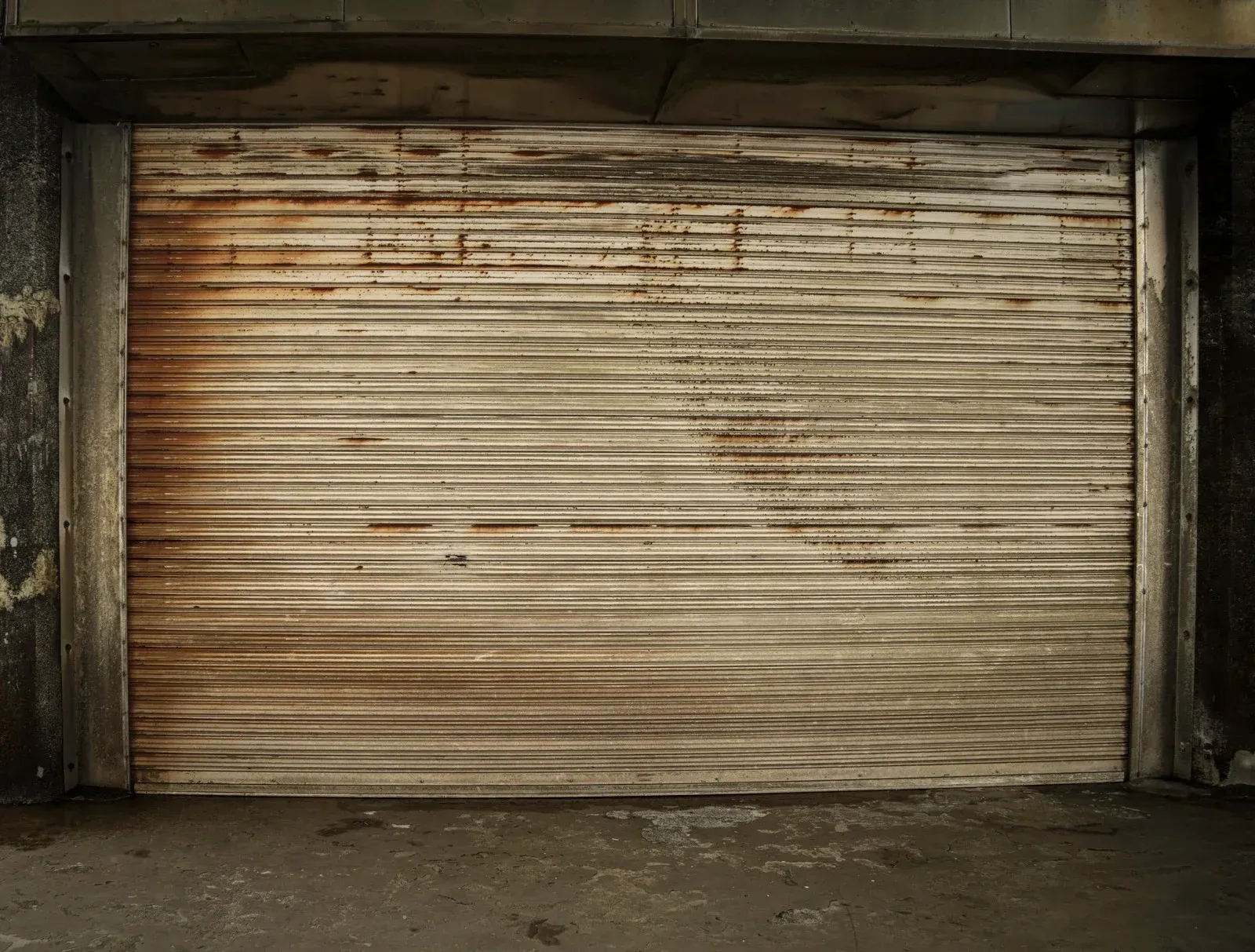 A weathered, closed metal roll-up garage door; gray and brown tones, dirty.