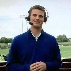 A man wearing headphones and a microphone is standing in front of a soccer field.