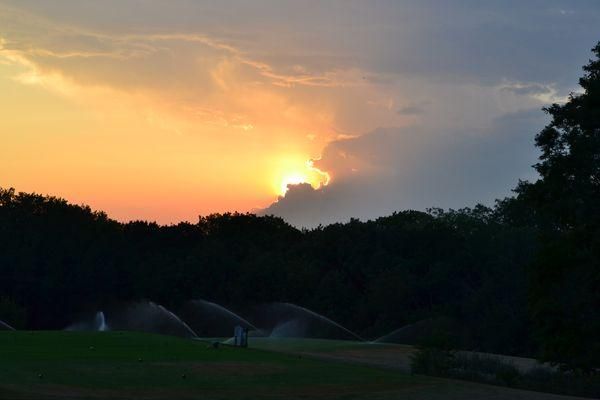 A sunset over a field with trees and sprinklers in the foreground