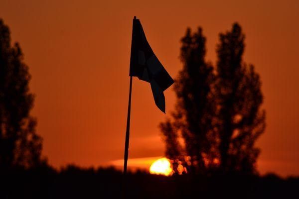 A golf flag is silhouetted against a sunset sky with trees in the foreground.