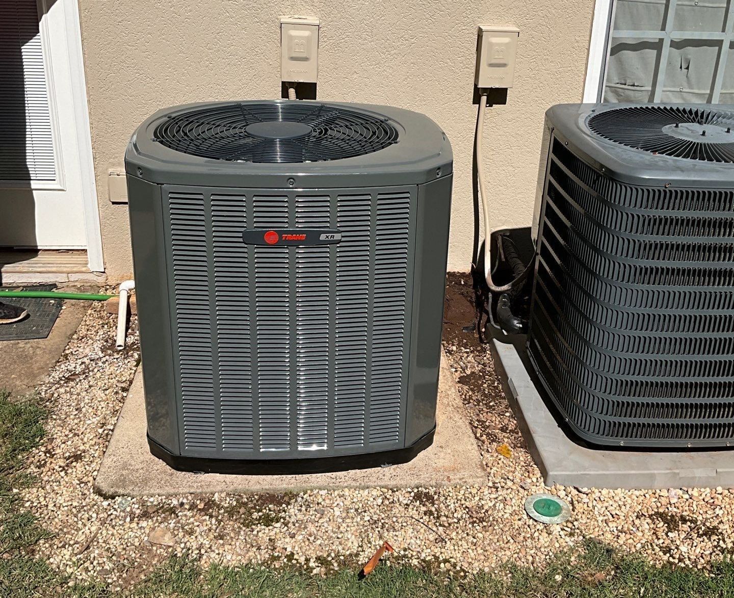 Two air conditioners are sitting next to each other on the side of a house.