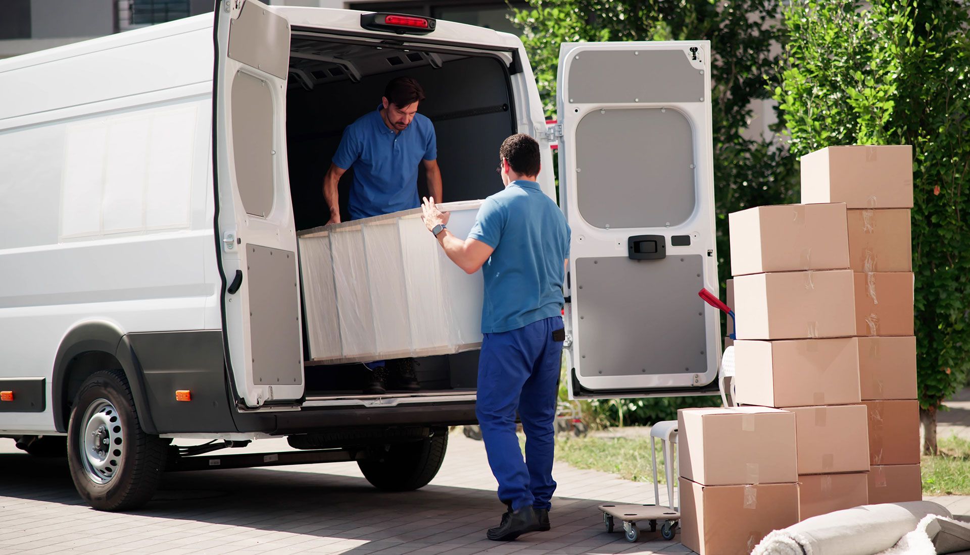 Two movers loading a large white item into a white van; boxes are stacked nearby.