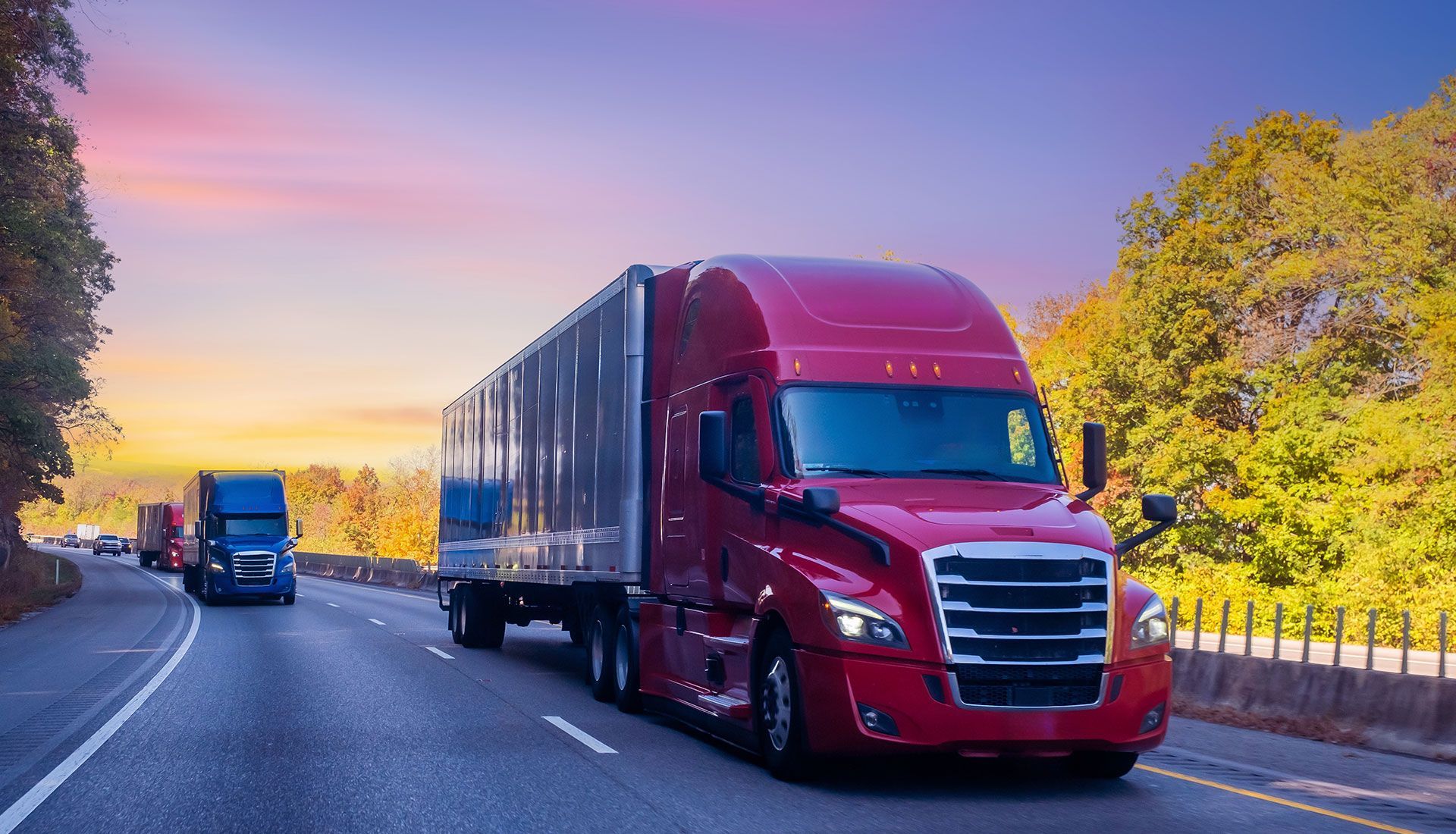 Red semi-truck driving on a highway with trees in the background under a sunset sky.