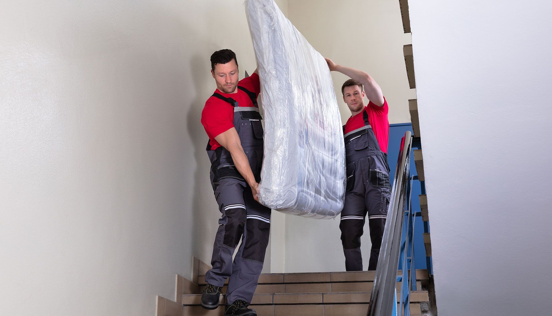 Two movers in red shirts and gray overalls carrying a mattress up stairs.