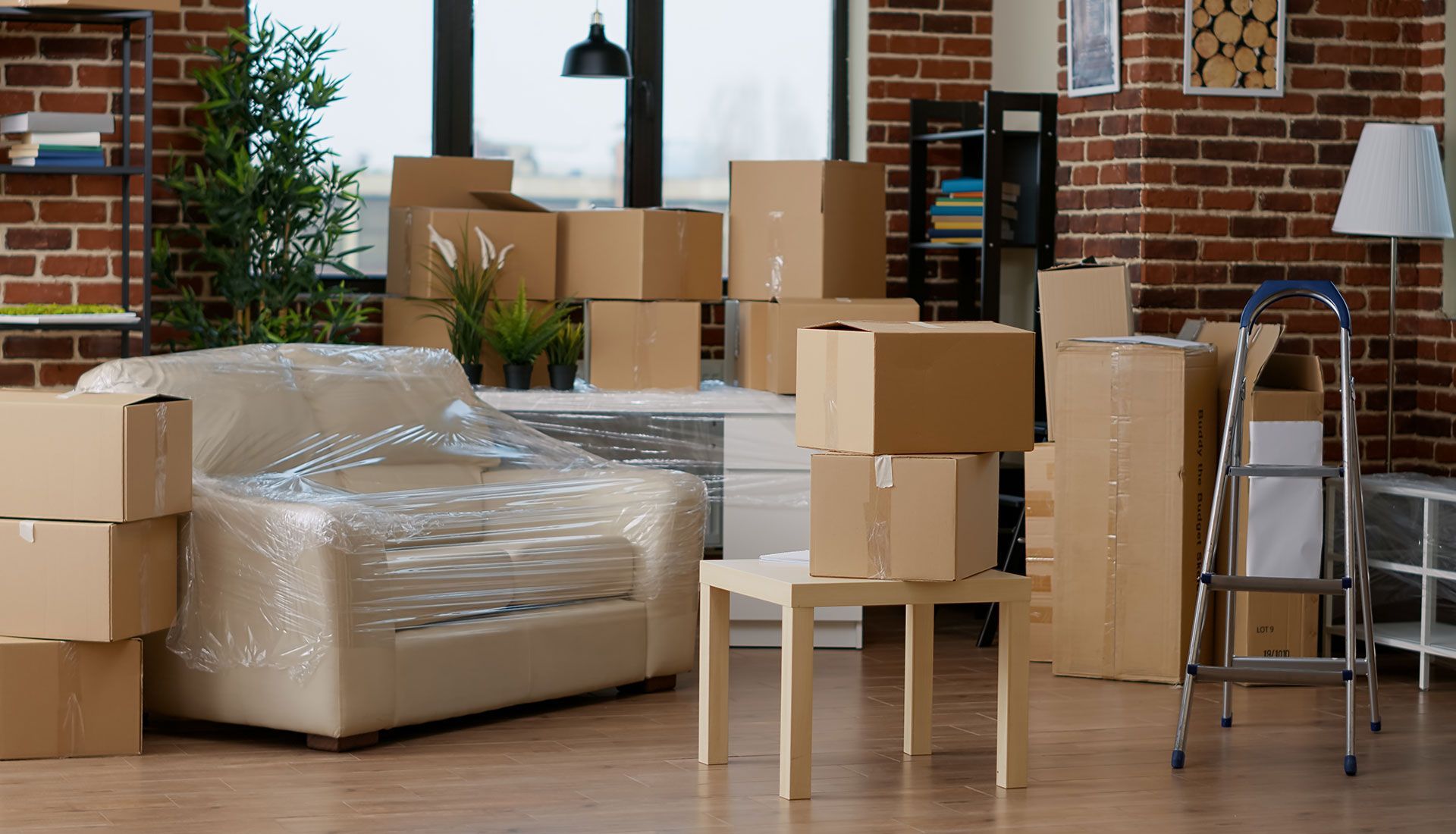 Boxes stacked in a room with packed furniture, ready for a move, next to a ladder and exposed brick wall.