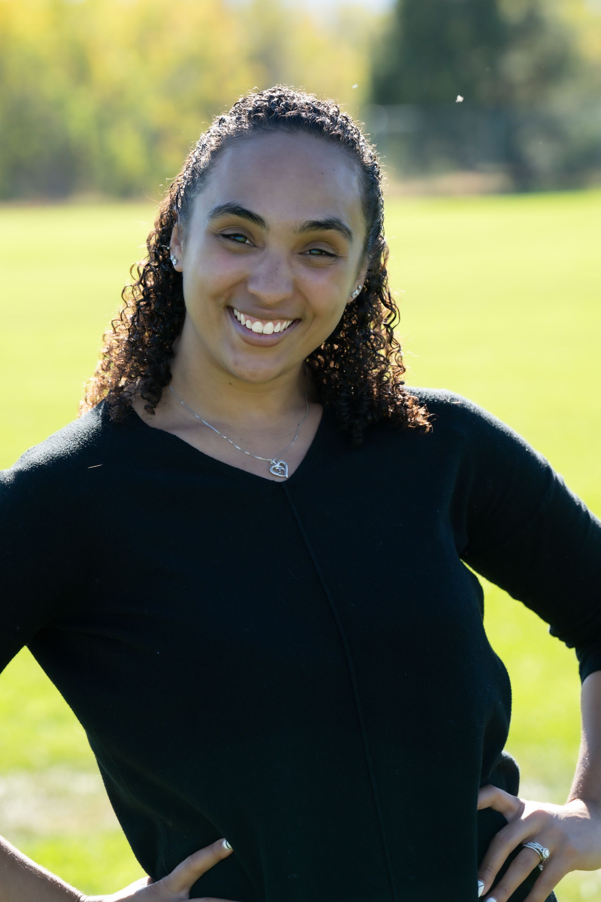 A woman in a black sweater is standing in a field with her hands on her hips.