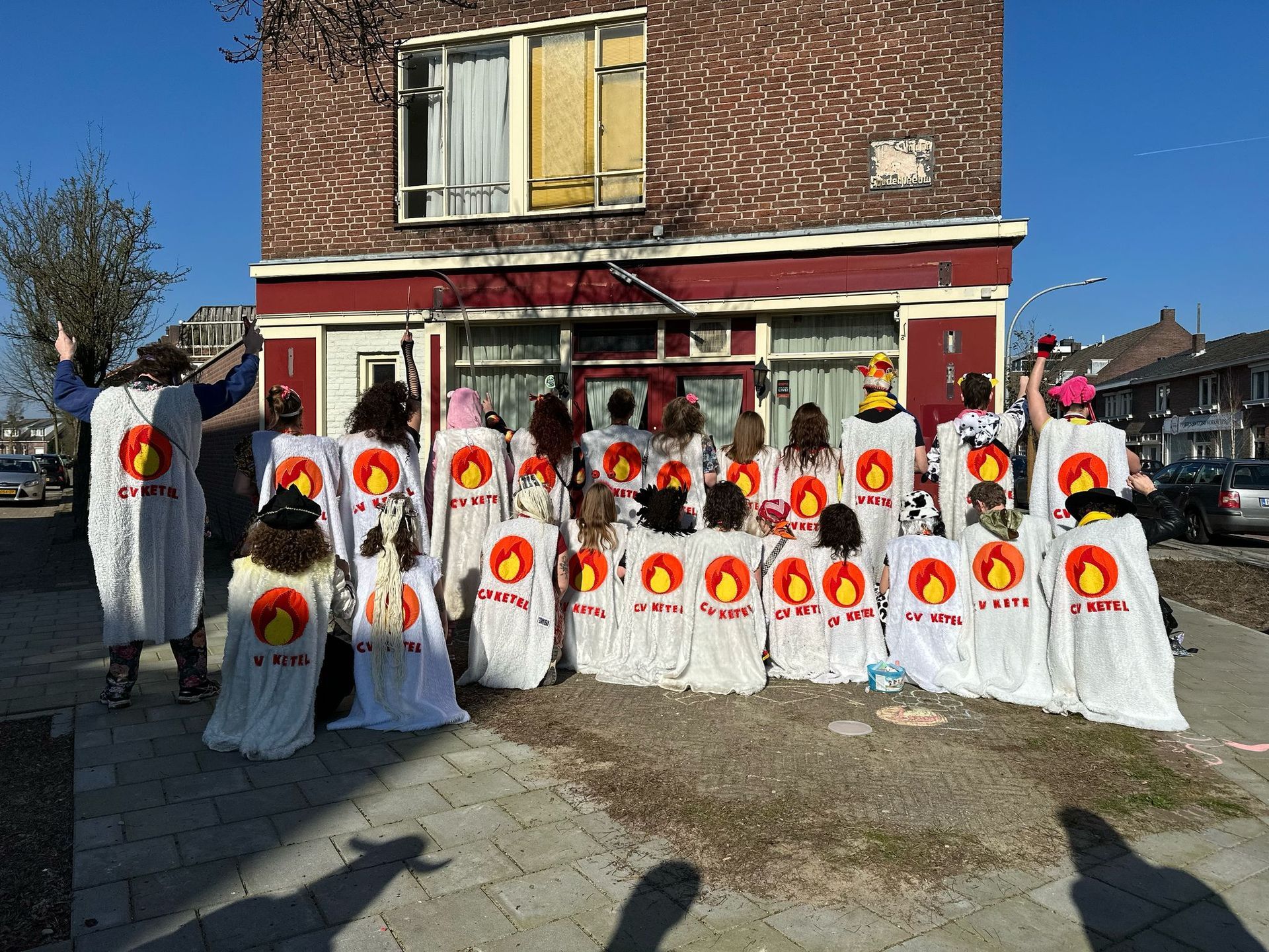 Een groep mensen in witte kostuums met een zonnesymbool poseert voor een bakstenen gebouw.