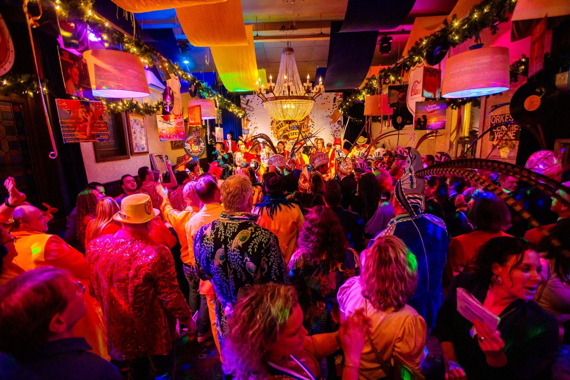 Feestje; volle ruimte met dansende mensen, kleurrijke verlichting en versieringen.