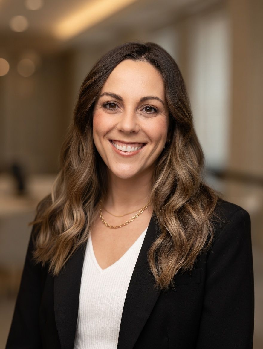 A professional headshot of a smiling person with long, wavy brown hair, wearing a white top under a black blazer.