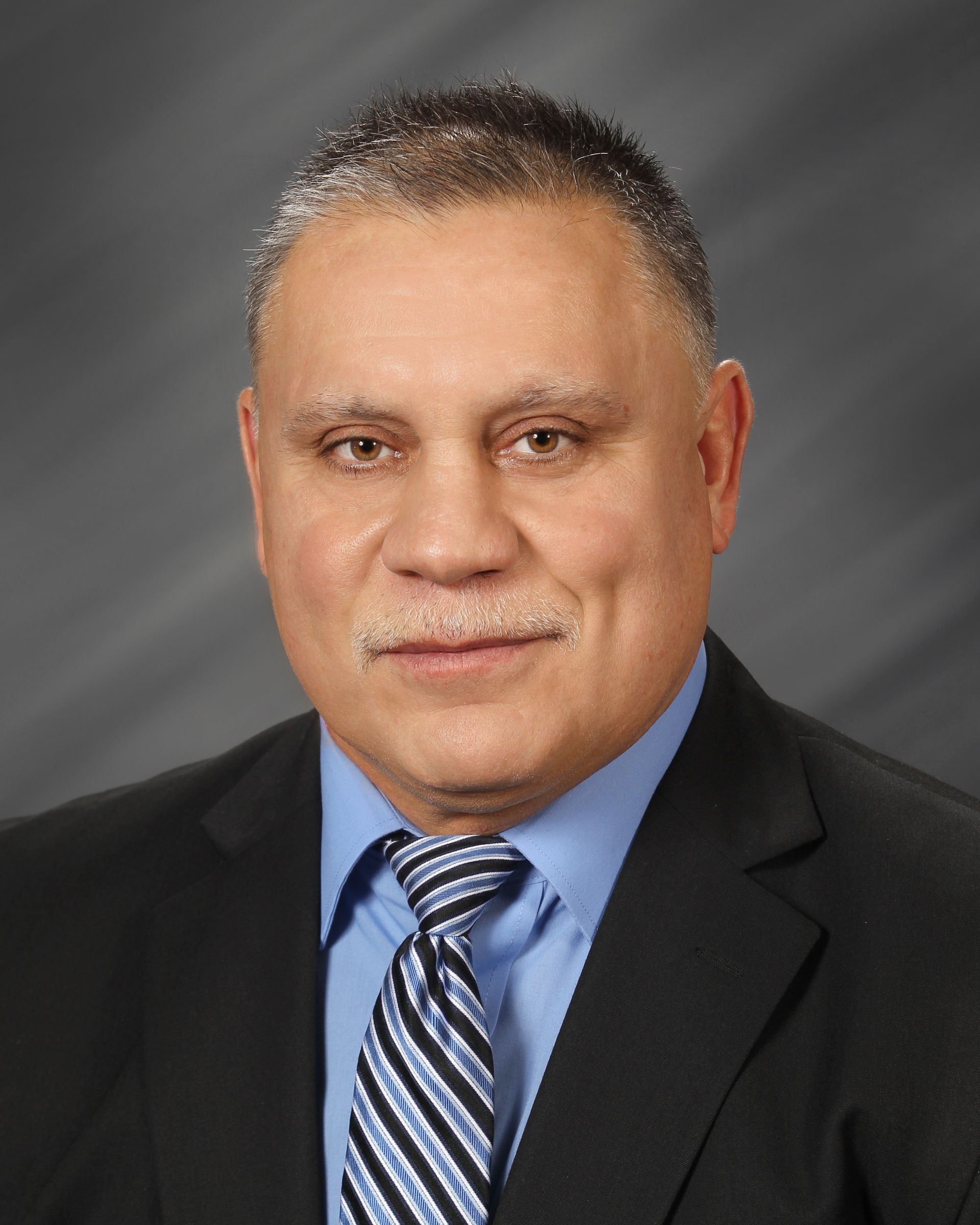 A man in a black suit, blue collared shirt, and striped tie against a gray background.