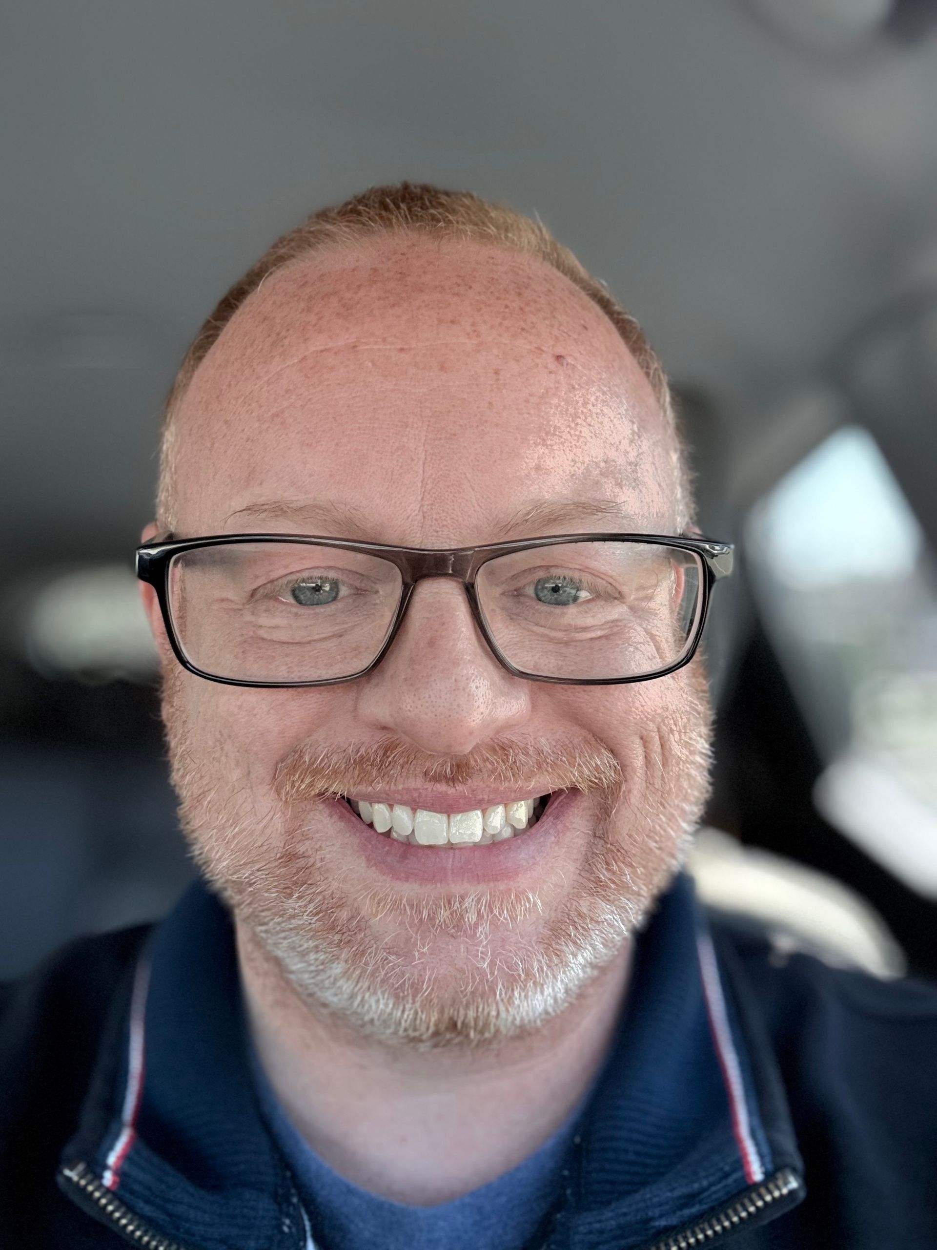 A smiling person with a light beard and glasses, centered in a portrait shot against a soft-focus background.
