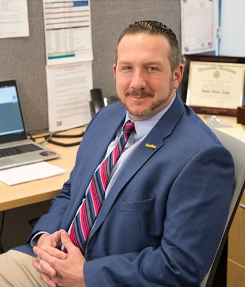 A professional wearing a blue suit and striped tie sits at a desk with a computer and framed certificate in an office.