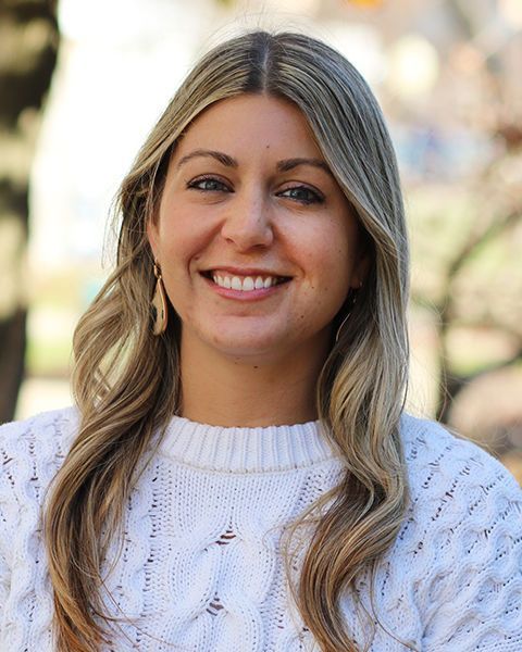 A smiling person with long blonde hair wearing a white cable-knit sweater and dangling earrings, outdoors.