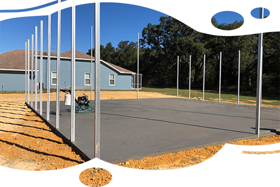 Construction site: metal poles erected on a gray concrete pad. A house is in the background under a blue sky.