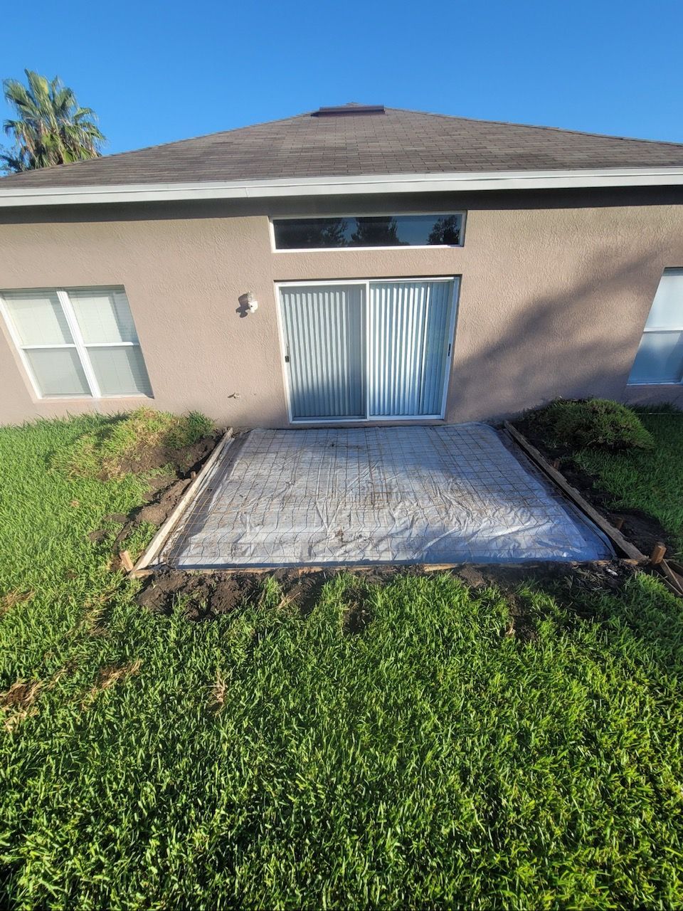 Concrete patio being built outside a house with sliding glass doors. Green grass surrounds it.