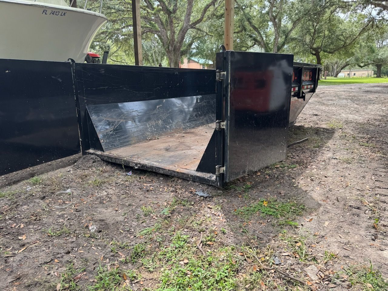 Black dumpster with open door on dirt ground, next to boat and trees.