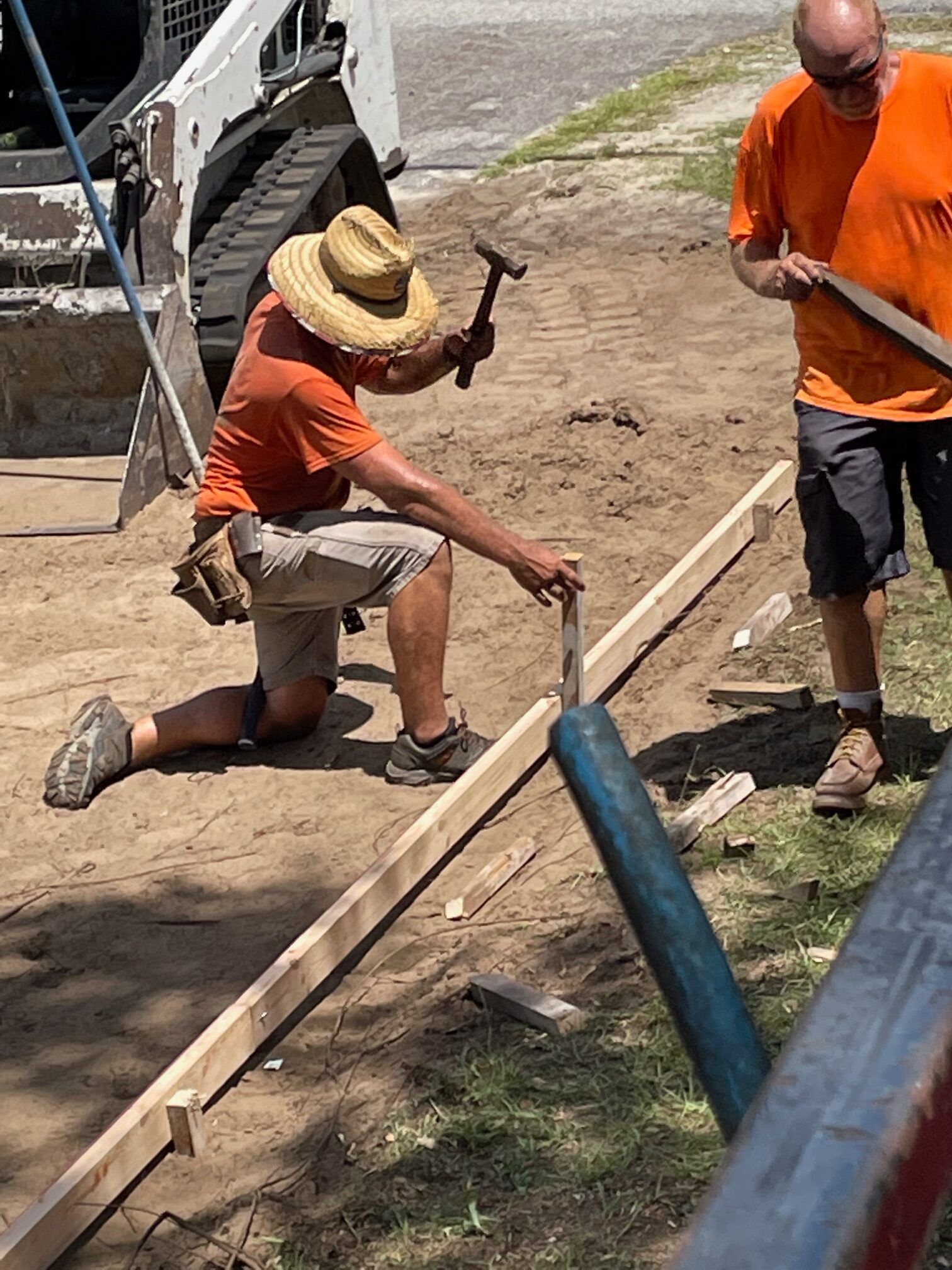Two construction workers building a frame; one kneels hammering a stake, the other holds a measuring stick.