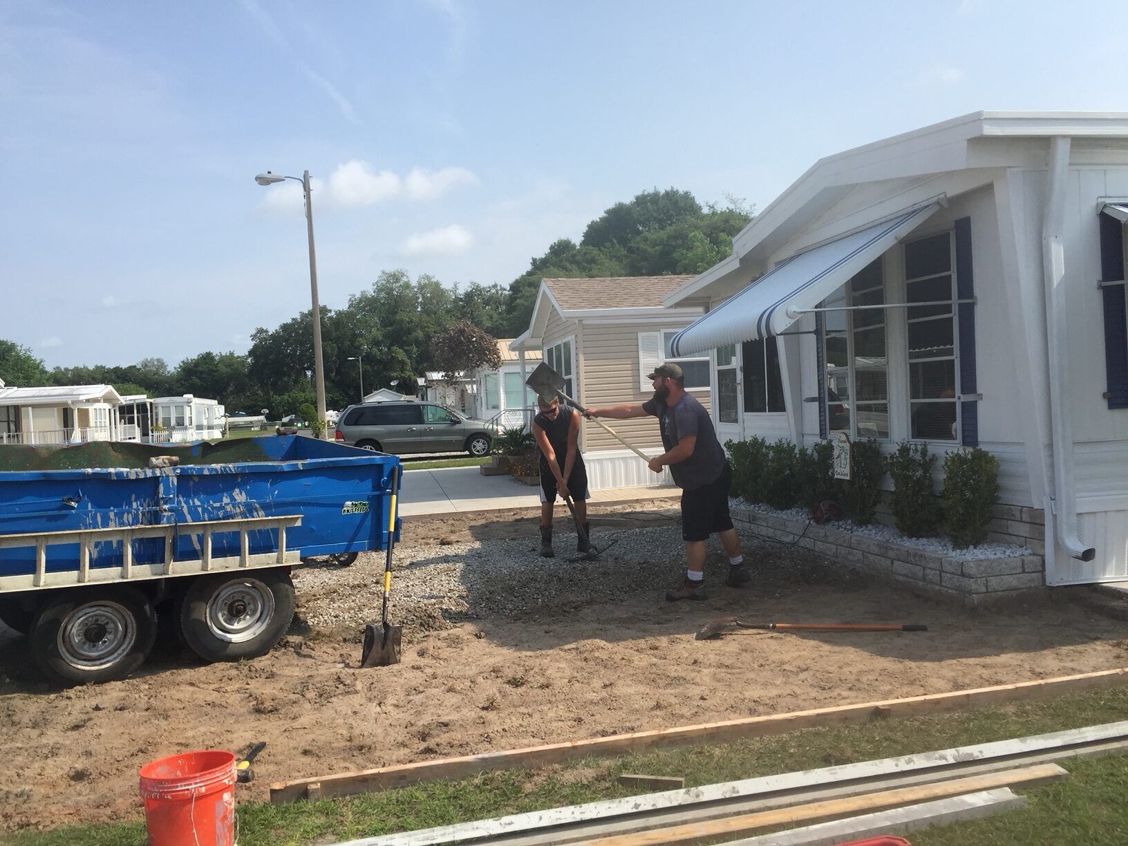 Two men working on landscaping in front of a mobile home, next to a trailer filled with gravel.