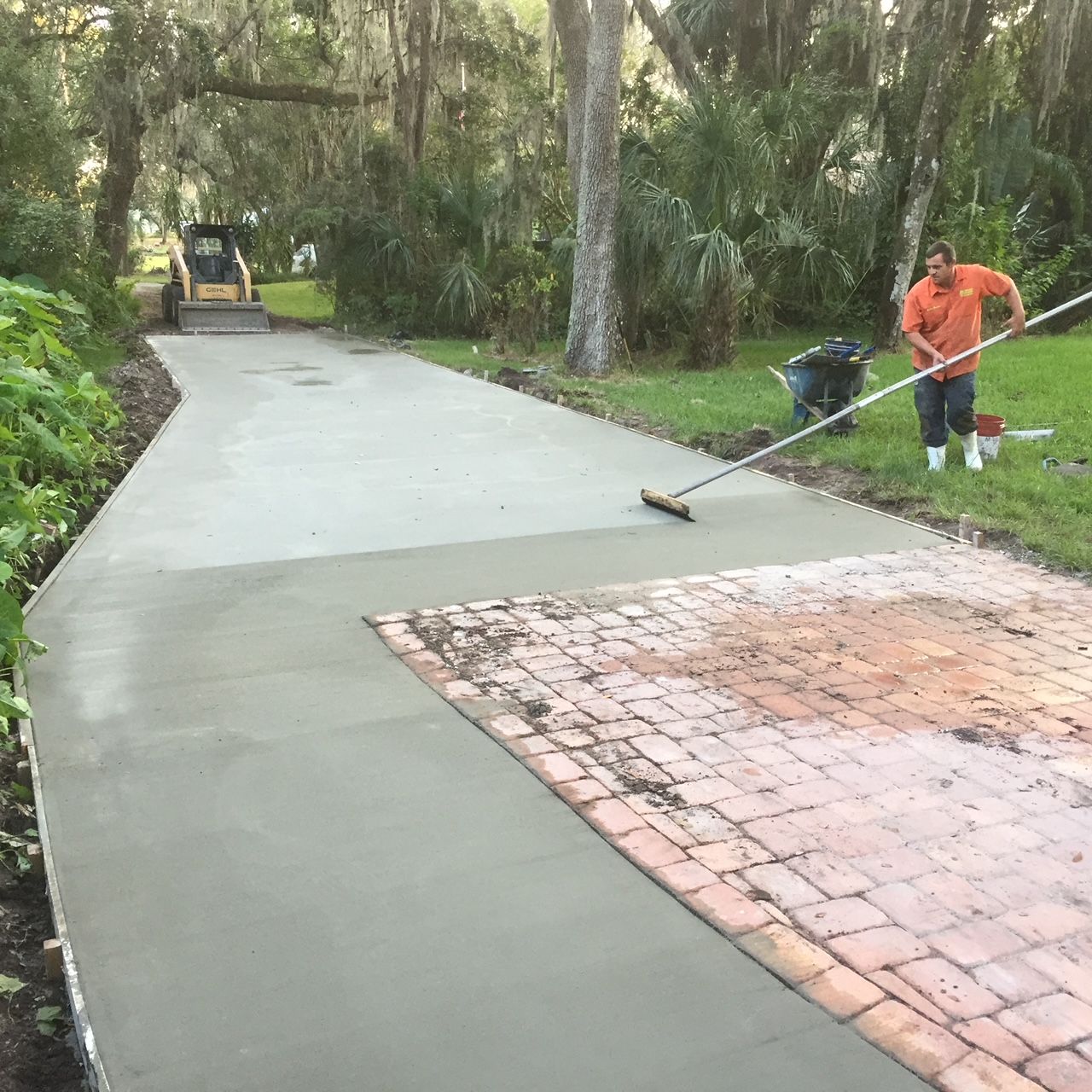 A worker smoothing fresh concrete on a driveway with brick pavers, trees in the background.