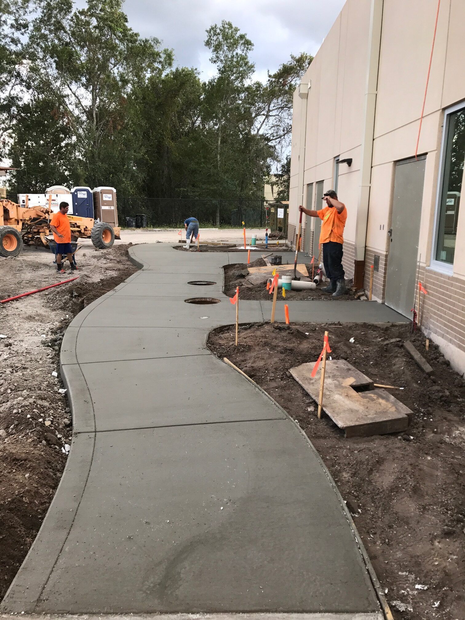 Workers pouring concrete sidewalk outside a building. One worker gestures; another operates equipment.