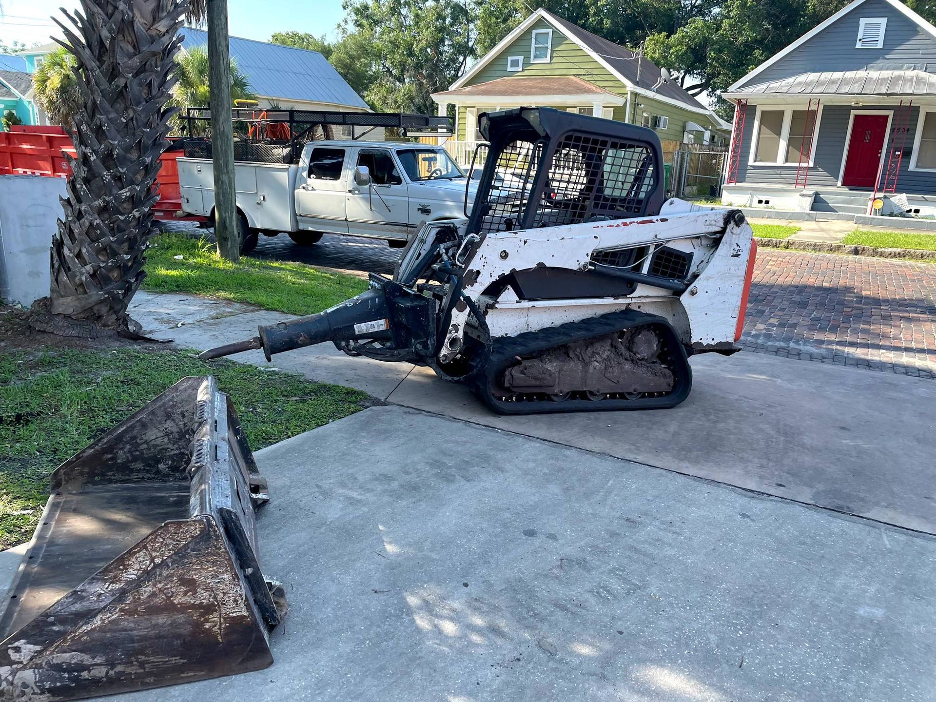 White Bobcat skid steer on a sidewalk with a pickup truck parked behind it, houses in background.