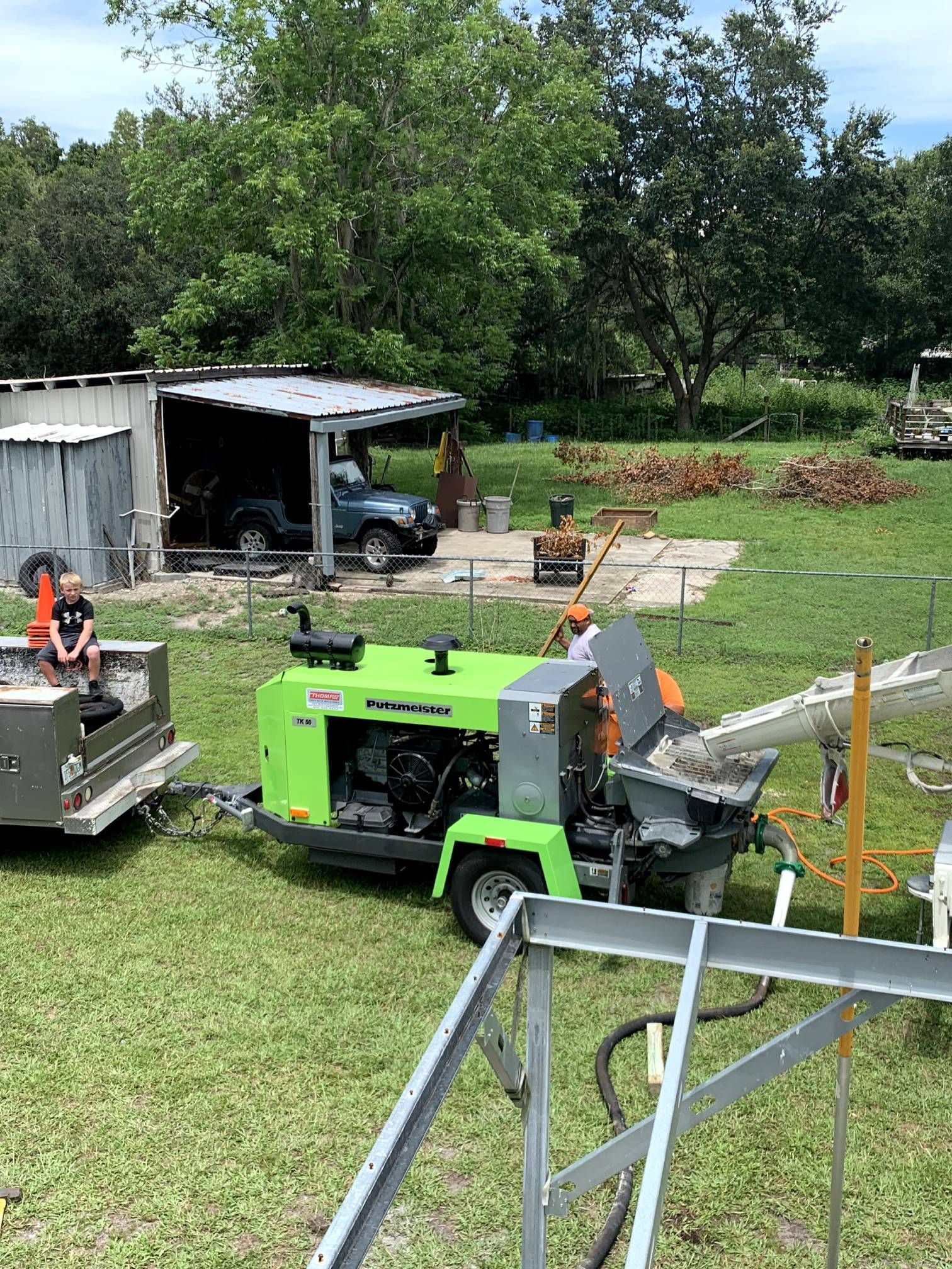 A green concrete pump truck pours concrete outdoors. A person guides the flow near a structure.