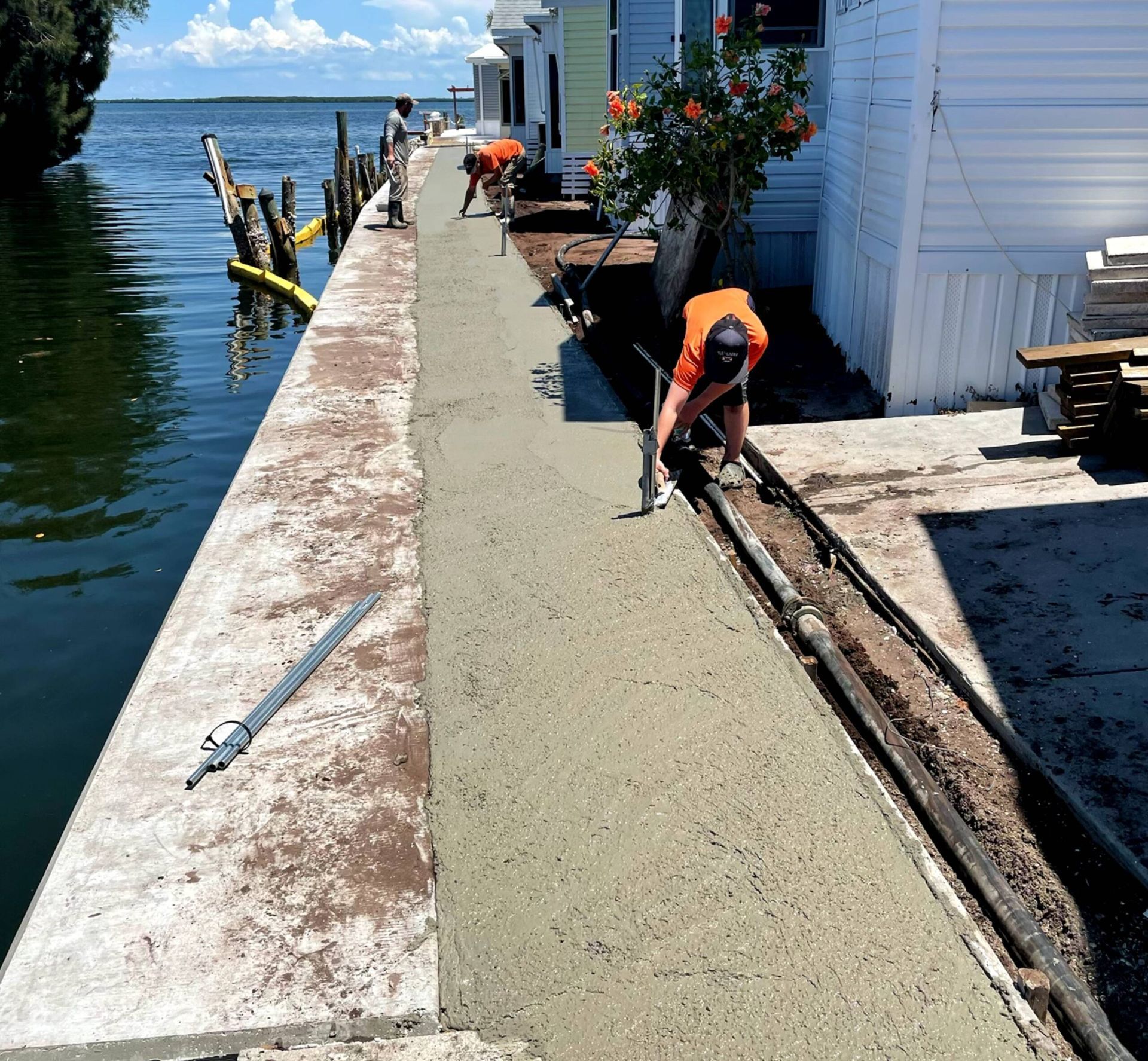 Construction workers laying concrete along a waterfront, next to homes.