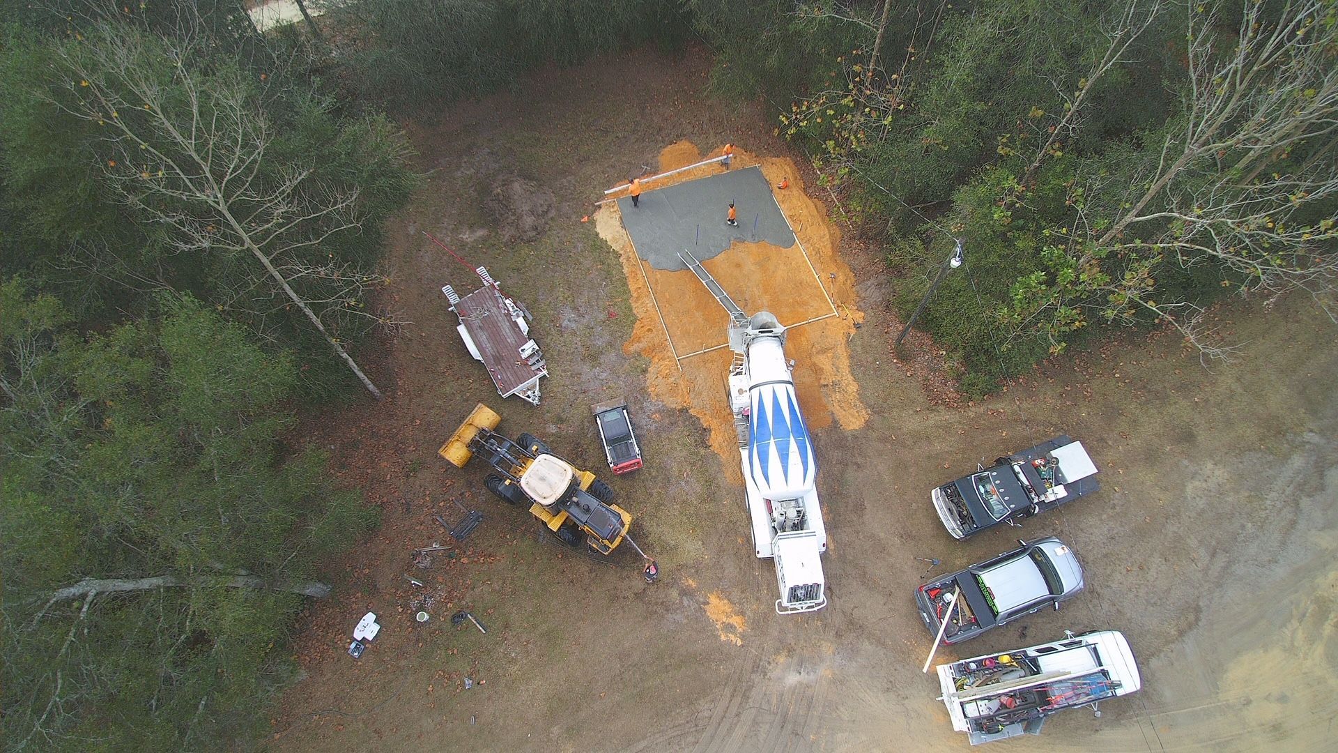Construction site, aerial view: concrete being poured.  Mixer truck, excavator, vehicles, and trees surround the work area.