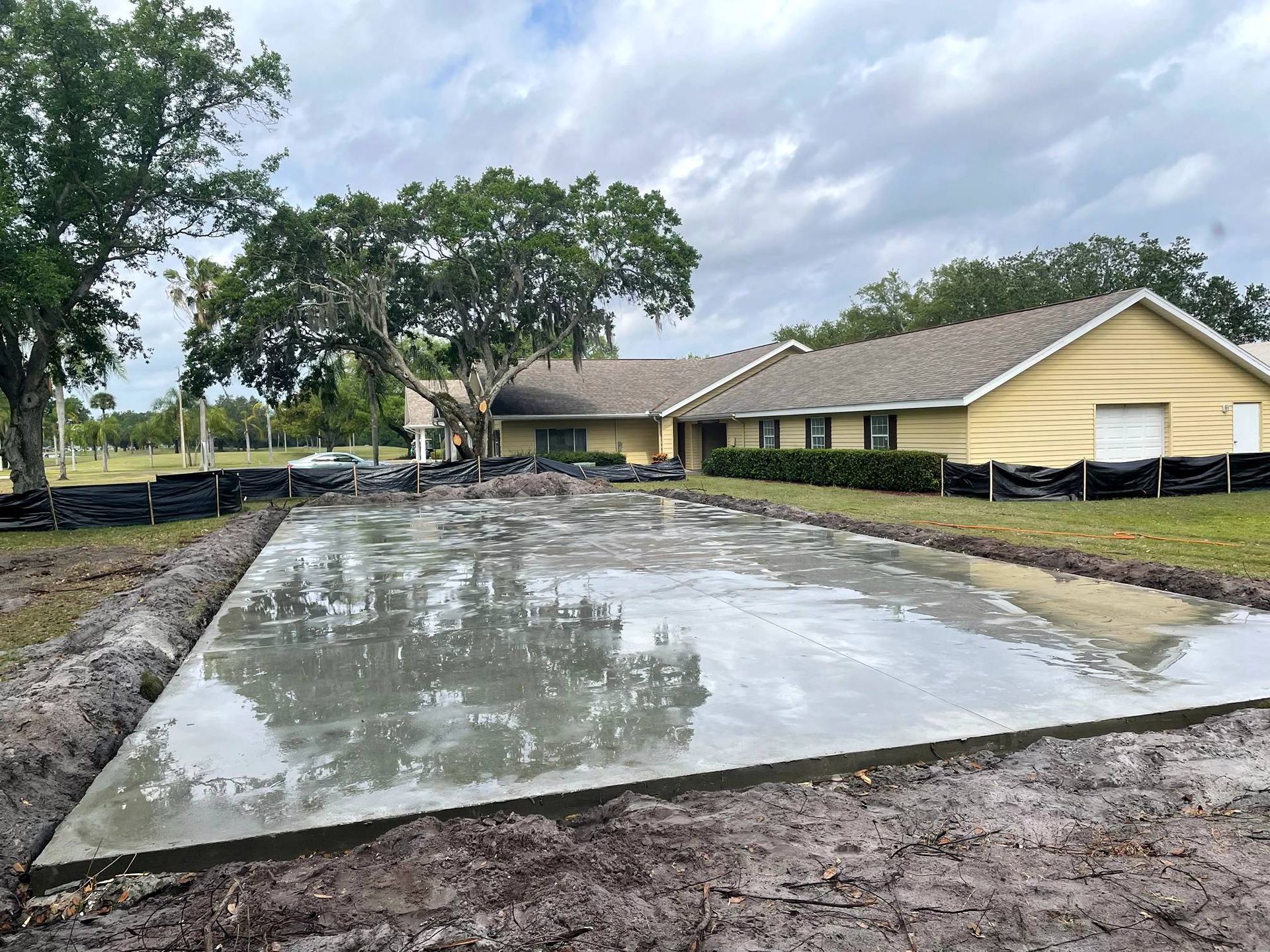 Newly poured concrete slab, next to a building, with surrounding dirt and barriers.