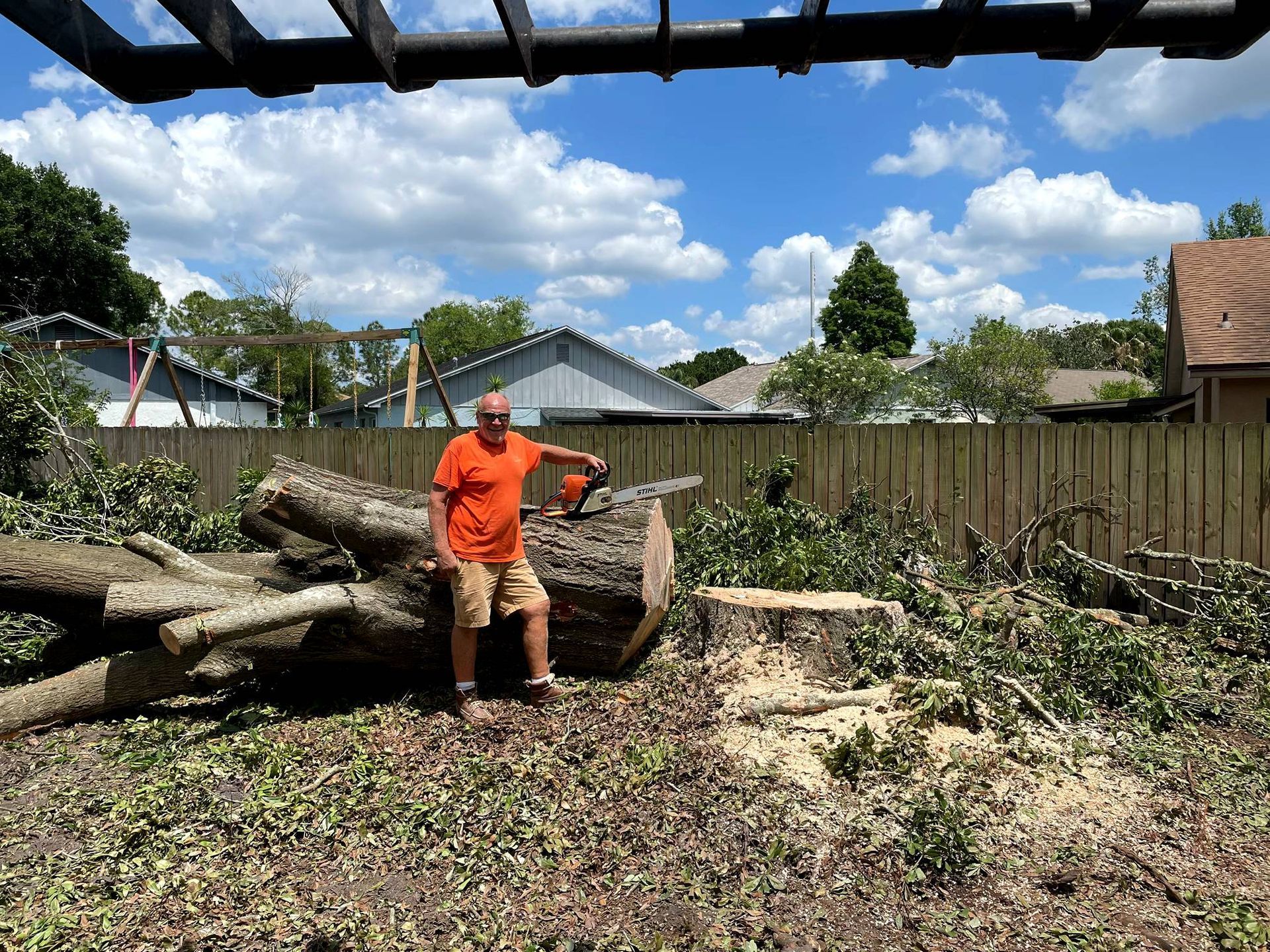 Man stands with a chainsaw in yard with cut tree. Sunny day, blue sky, houses and fence in background.