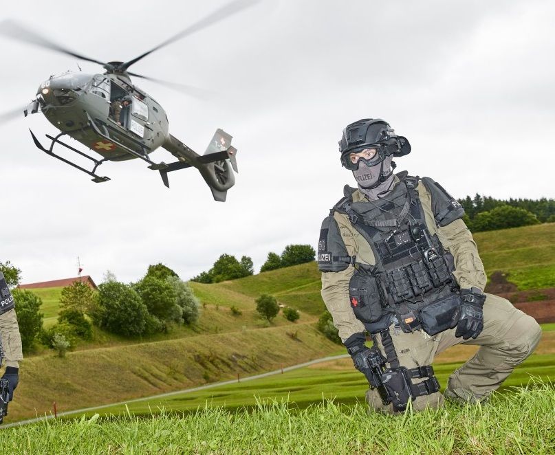 Ein Militärhubschrauber schwebt über einem getarnten Polizisten, der auf einem grasbewachsenen Feld kniet.