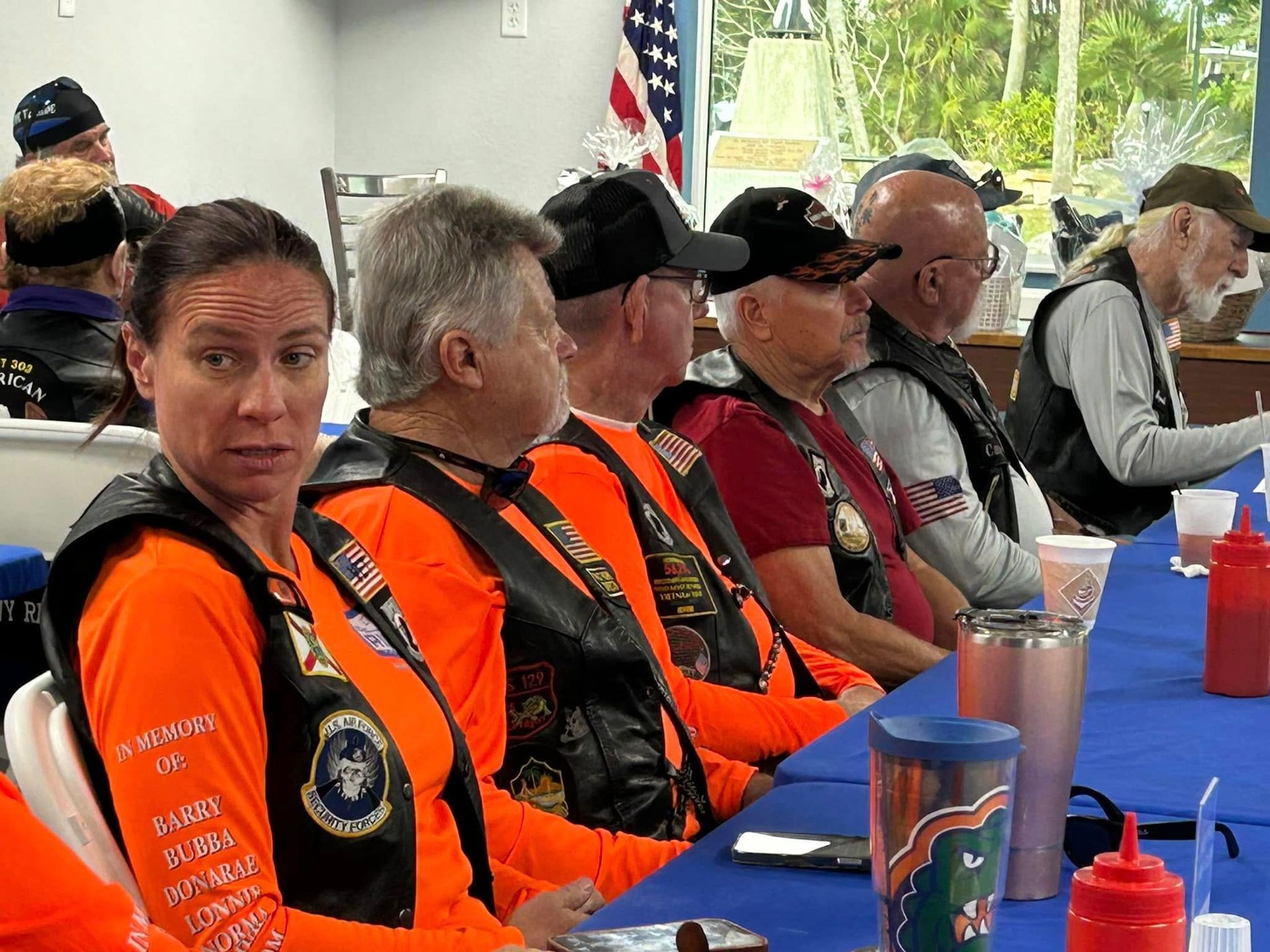 Group of people in orange vests at a table. American flag and interior setting are visible.