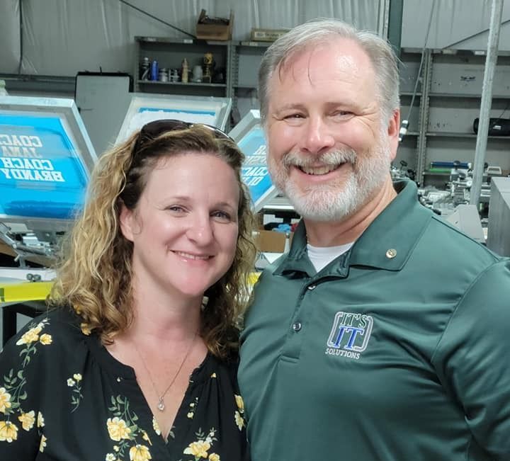 A woman and a man smiling in a workshop. The man wears a green shirt. Behind them are printing screens.
