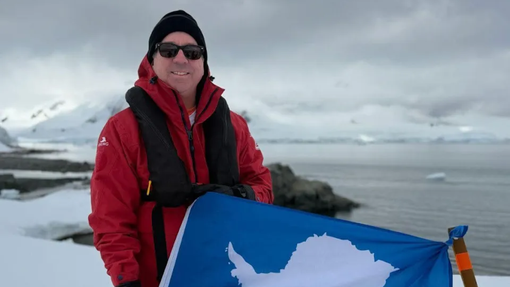 Man in red jacket and sunglasses holds Antarctic flag, posing in snowy landscape near the ocean.