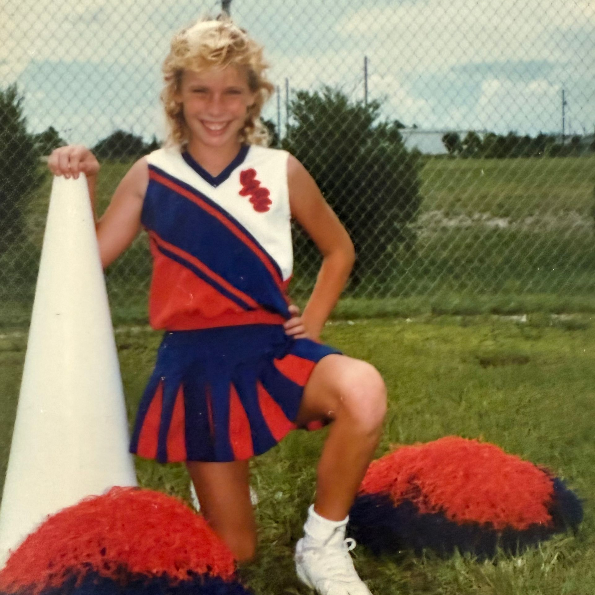 Blonde cheerleader kneels on grass, holding a megaphone and smiling. She wears a red, white, and blue uniform.