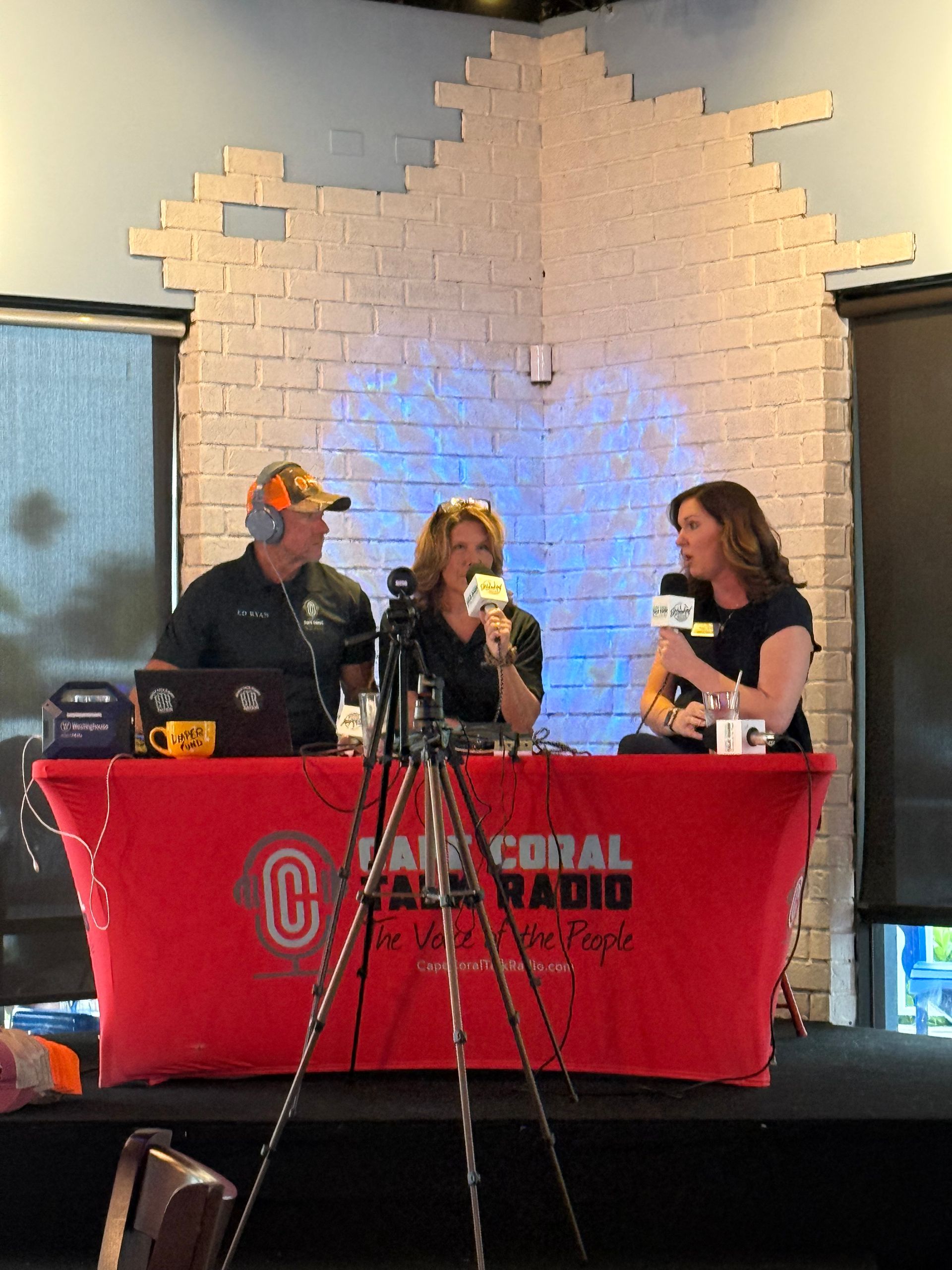 Three people at a table with microphones; broadcasting for Cape Coral Radio, in front of a brick wall.