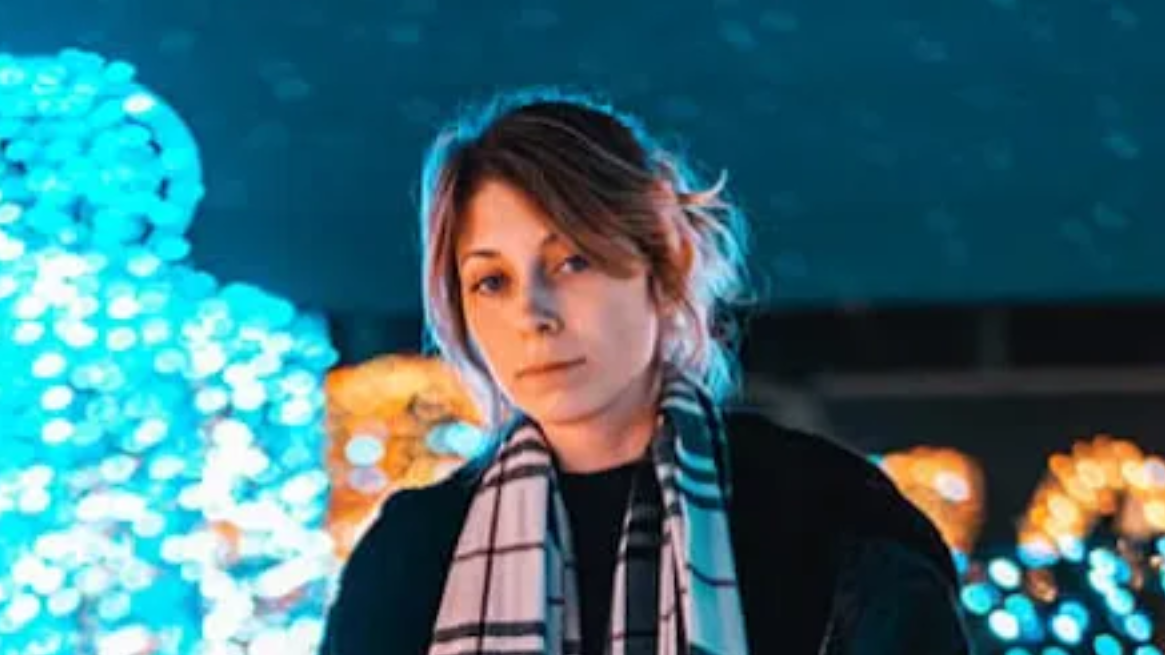 Woman with short hair and scarf, stands in front of blue and yellow lights.