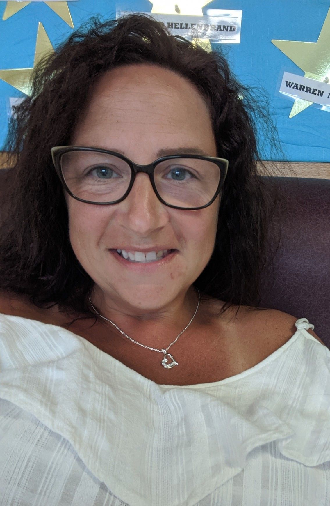 Woman with glasses smiles, wearing white top and silver necklace. Dark hair, blue background.