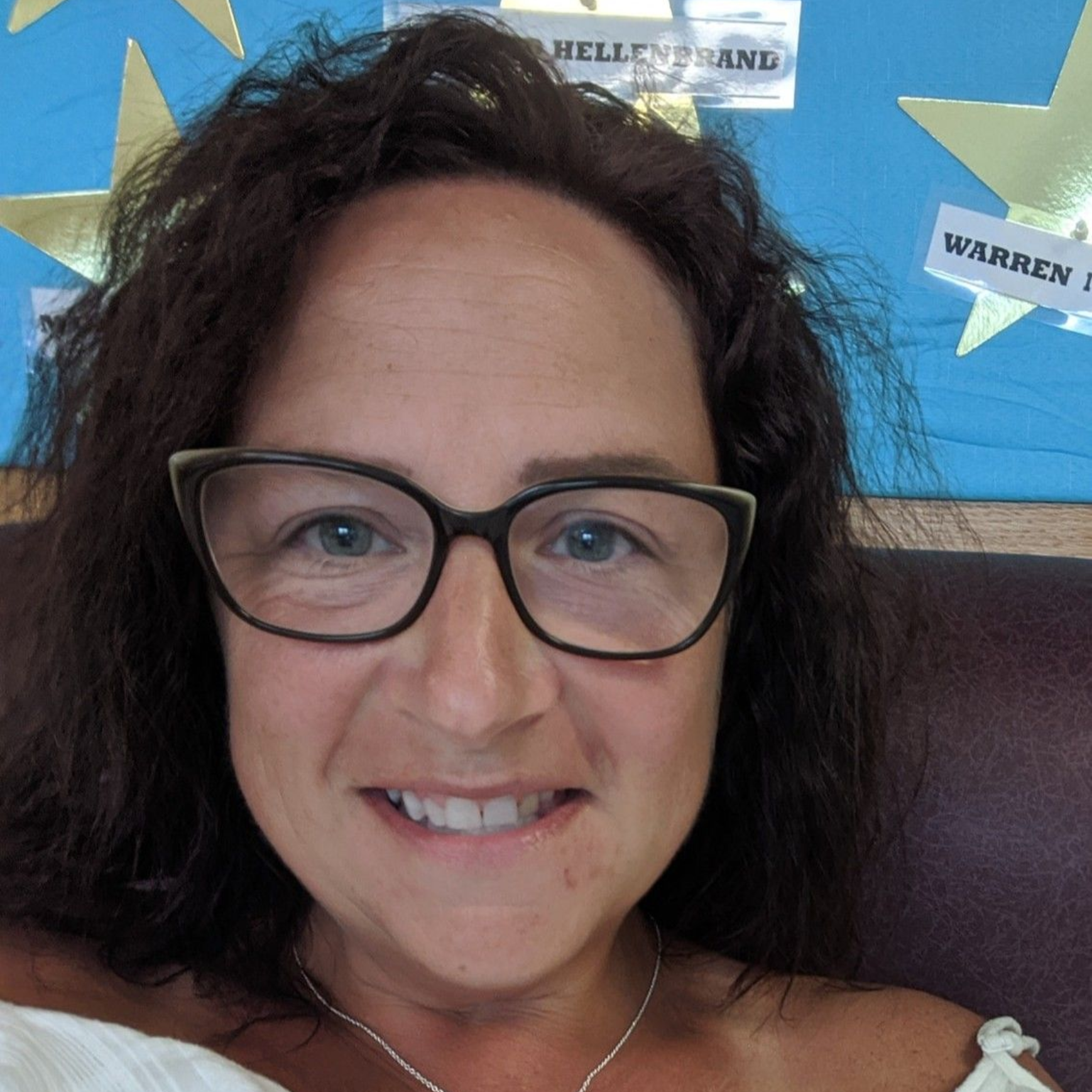 Woman with glasses smiles, wearing white top and silver necklace. Dark hair, blue background.