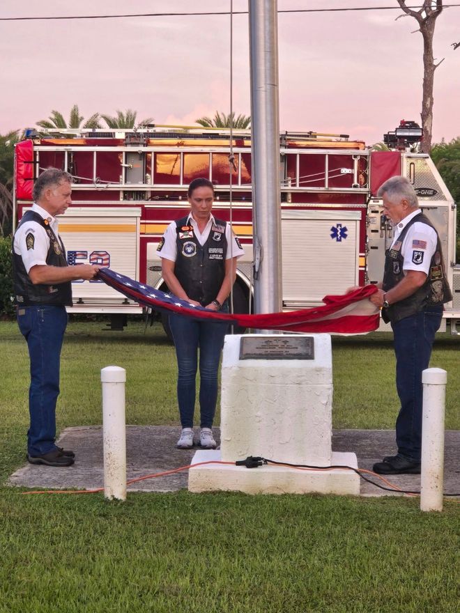 Three people folding an American flag at a flagpole, with a fire truck in the background.