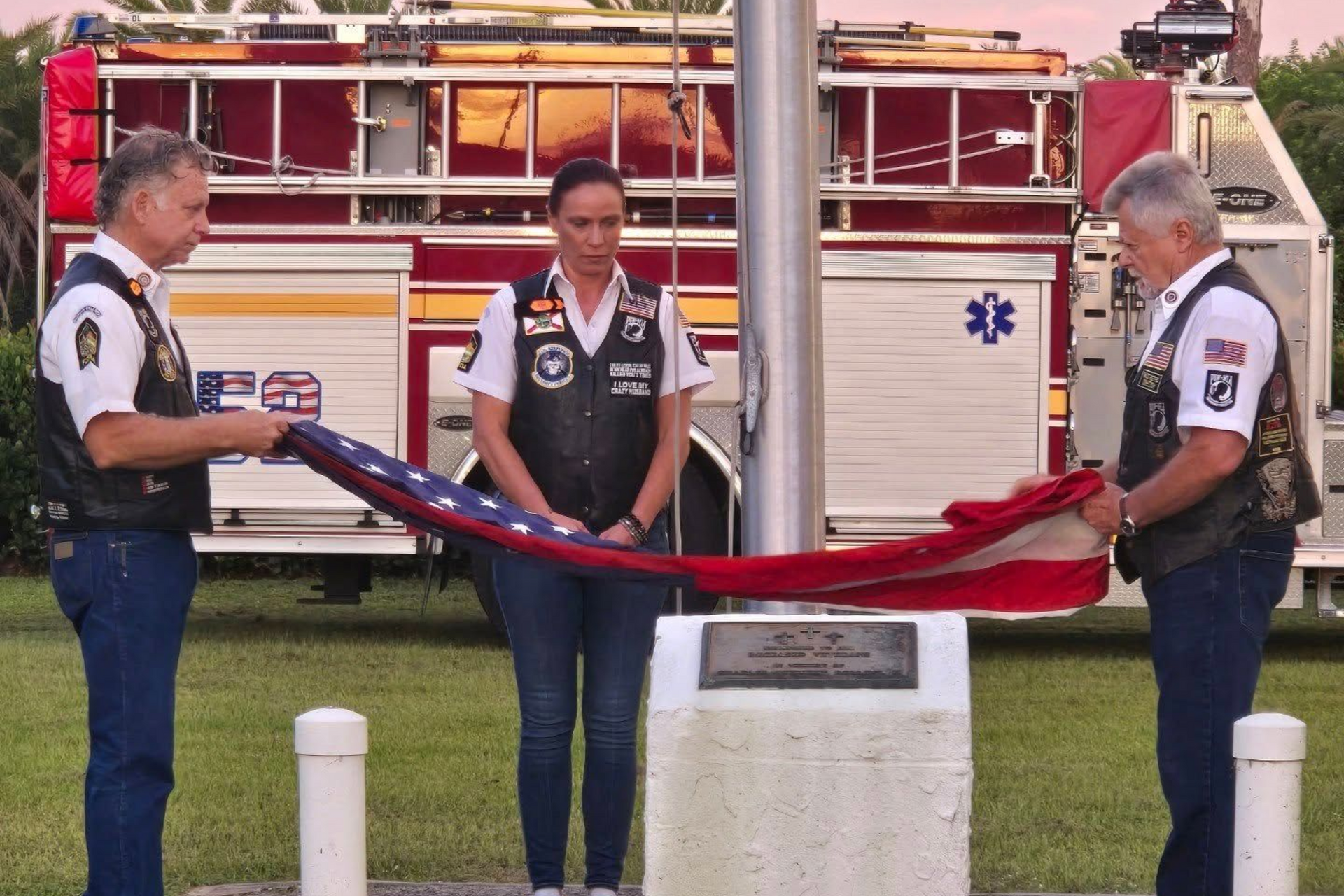 Three people folding an American flag at a flagpole, with a fire truck in the background.