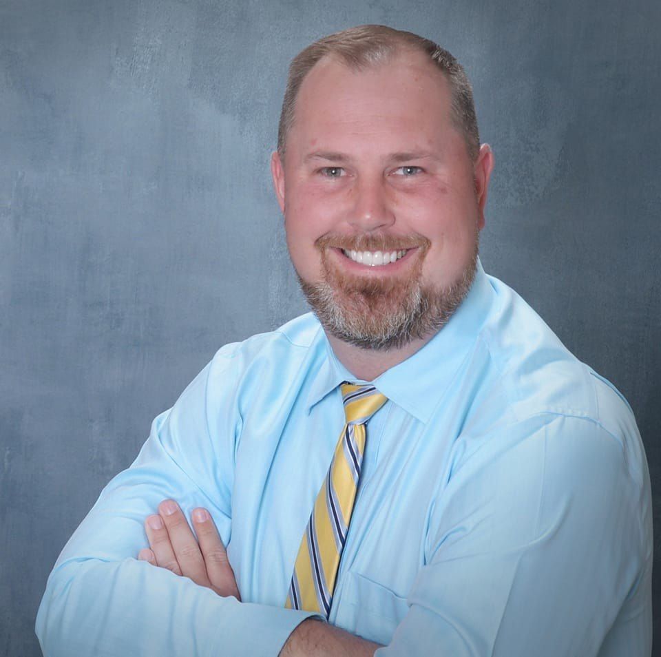 Man with beard, arms crossed, smiling, wearing light blue shirt and yellow striped tie.