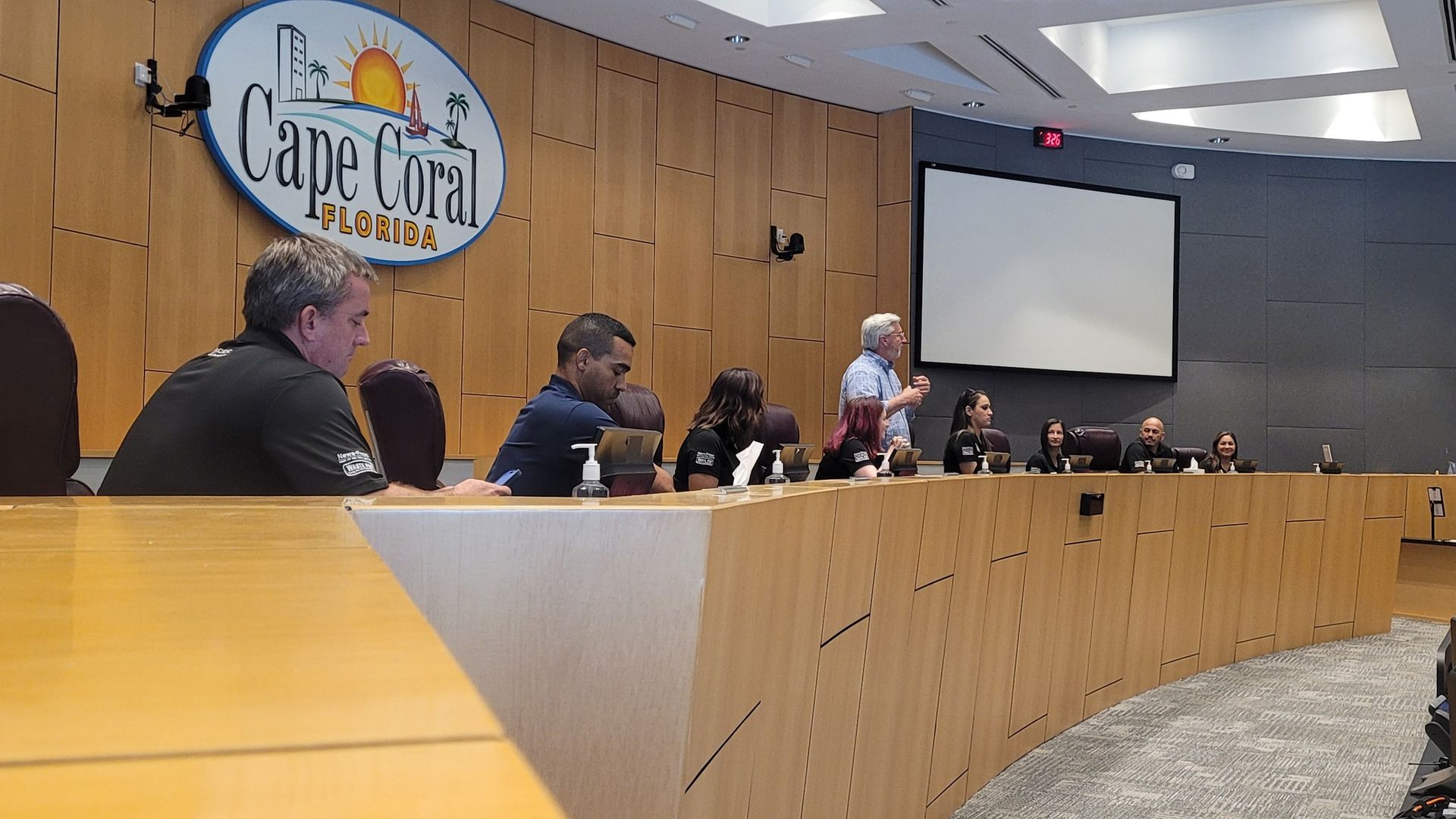 City council meeting in Cape Coral, Florida. People seated at a curved wooden dais, one speaking with a presentation screen.