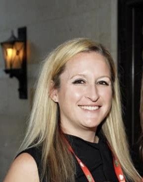 Blonde woman smiling, wearing a black top, standing indoors.
