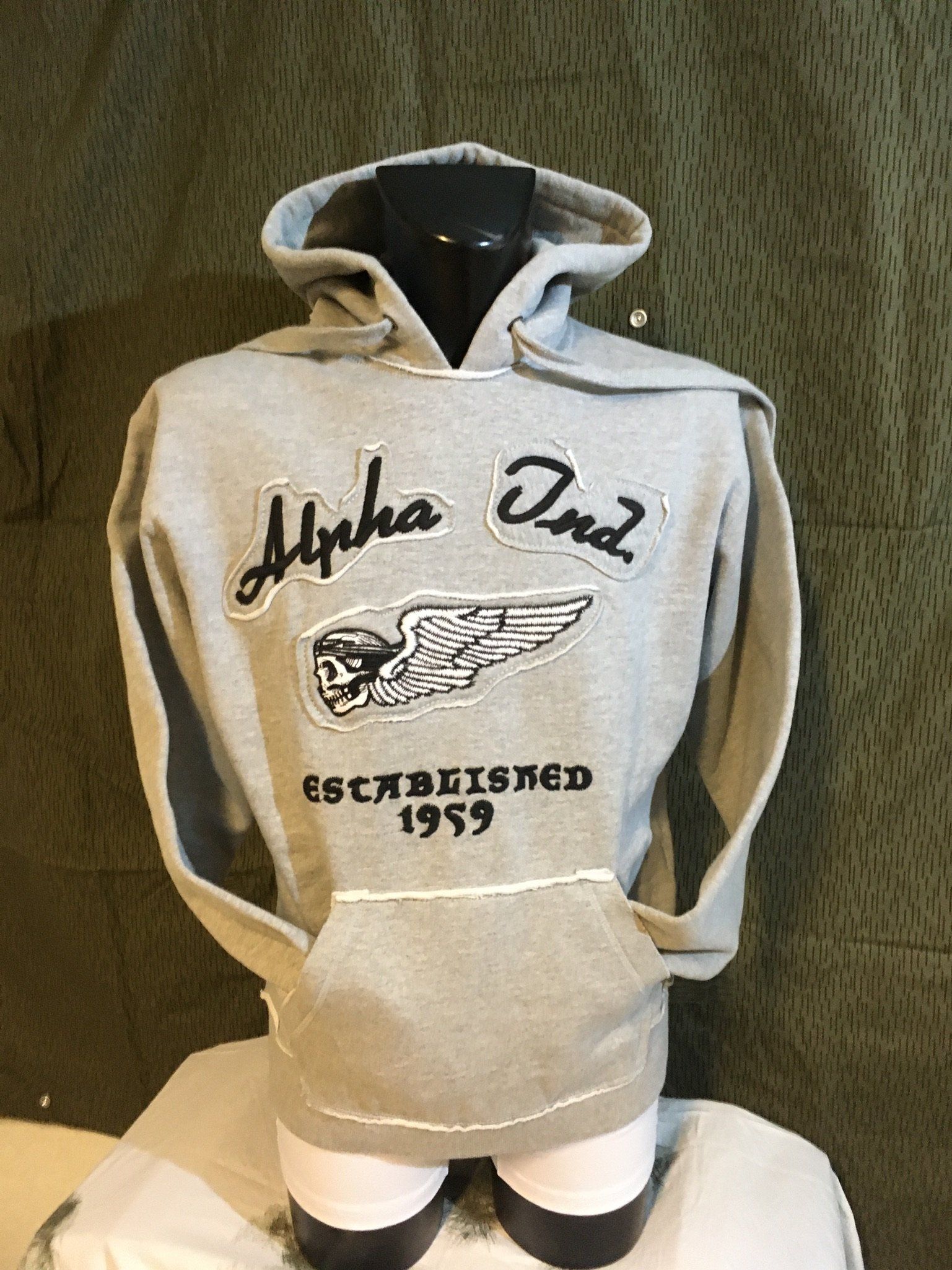 Graues Kapuzensweatshirt mit „Alpha Ond“ und einem geflügelten Emblem; gegründet 1959.