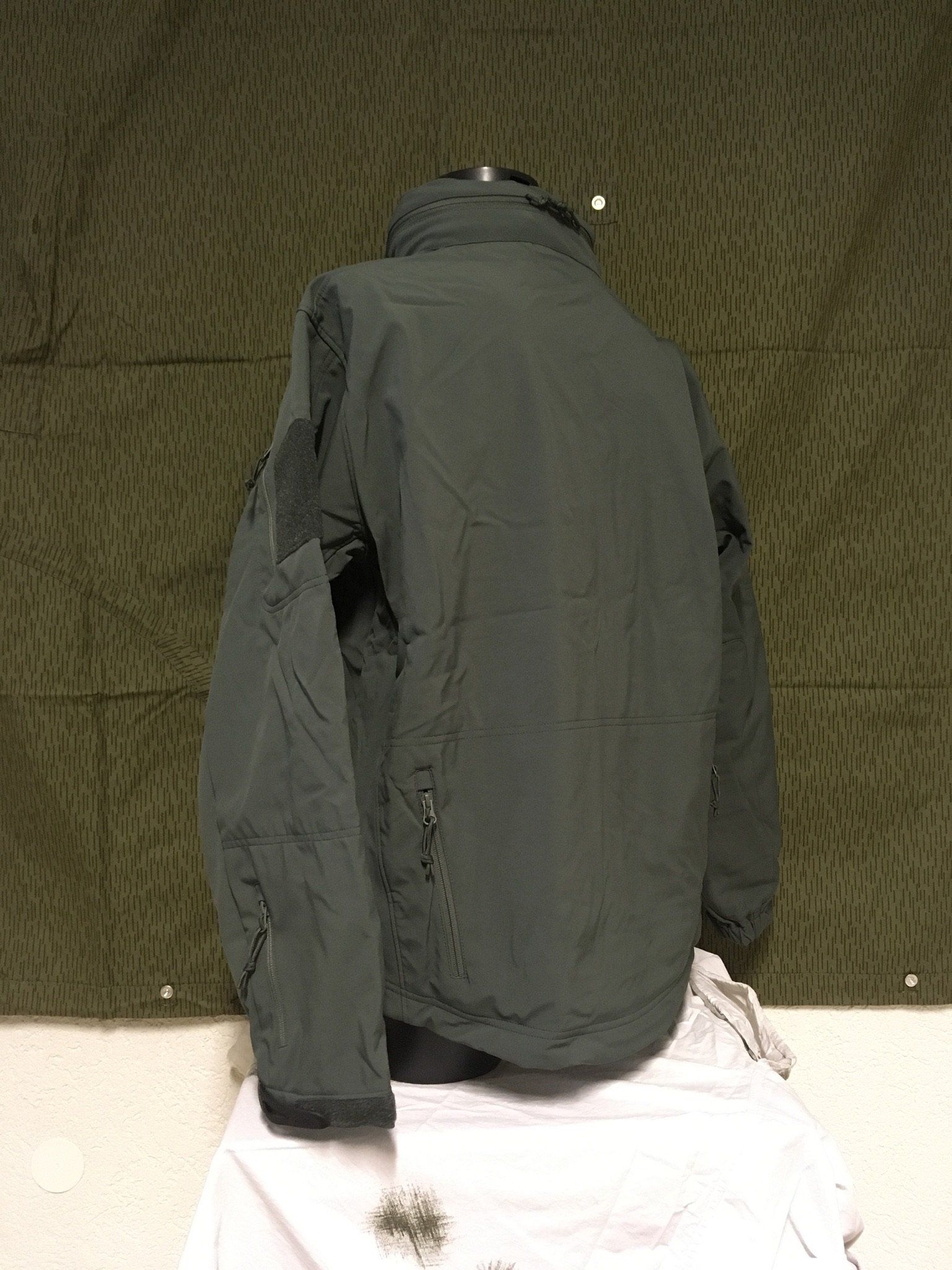 Dunkelgrüne Jacke, die auf einer weißen Fläche vor einem gemusterten grünen Hintergrund hängt.